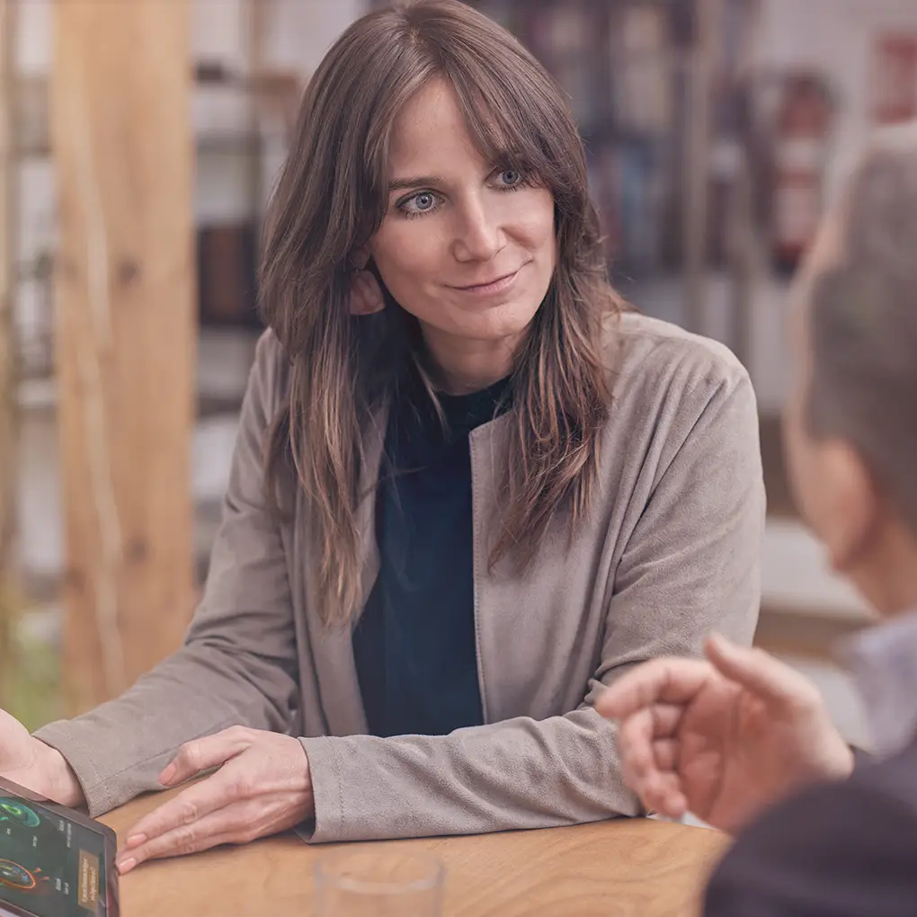Eine Frau sitzt an einem Holztisch, hält ein Tablet in der Hand und hört einer gegenüber sitzenden Person aufmerksam zu